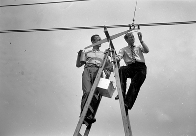 839988 Afbeelding van twee technici van N.S. tijdens het controleren van de meetapparatuur aan de bovenleiding voor ...